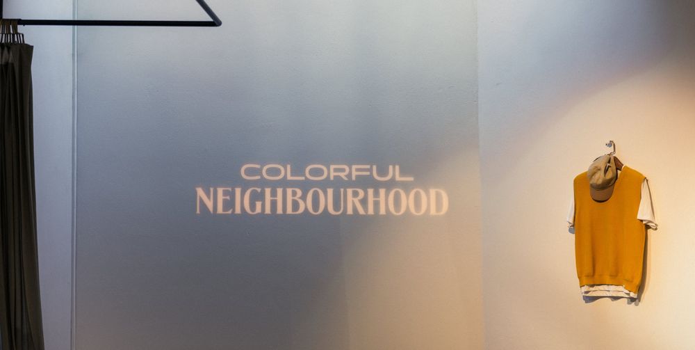 Schriftzug Colorful Neighbourhood an einer hellen Wand, daneben ein senfgelber Pullunder